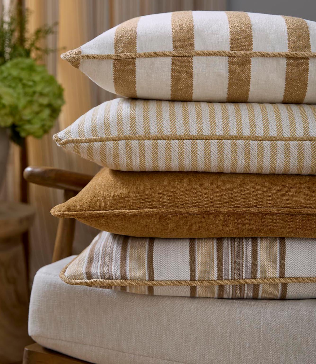 Stack of upholstered cushions in coordinating woven fabrics, combining plain and striped designs in warm ochre and soft neutral tones, creating a layered, relaxed look.