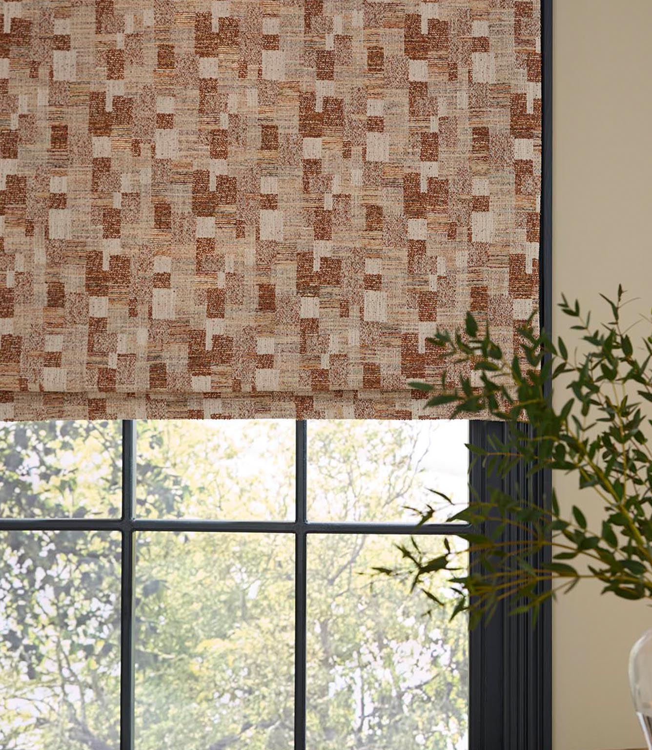 Roman blind in a woven geometric fabric with an abstract check pattern in warm rust and natural linen tones, styled over a black-framed window with soft daylight and greenery visible outside.