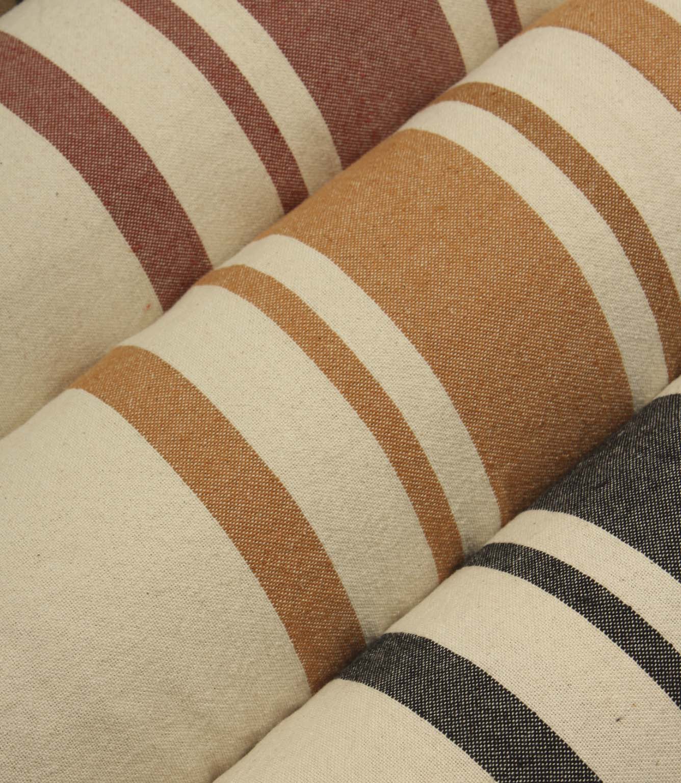 Woven striped furnishing fabrics in coral red, camel brown, and neutral beige tones, displayed as rolled fabric bolts with classic wide stripe detailing.