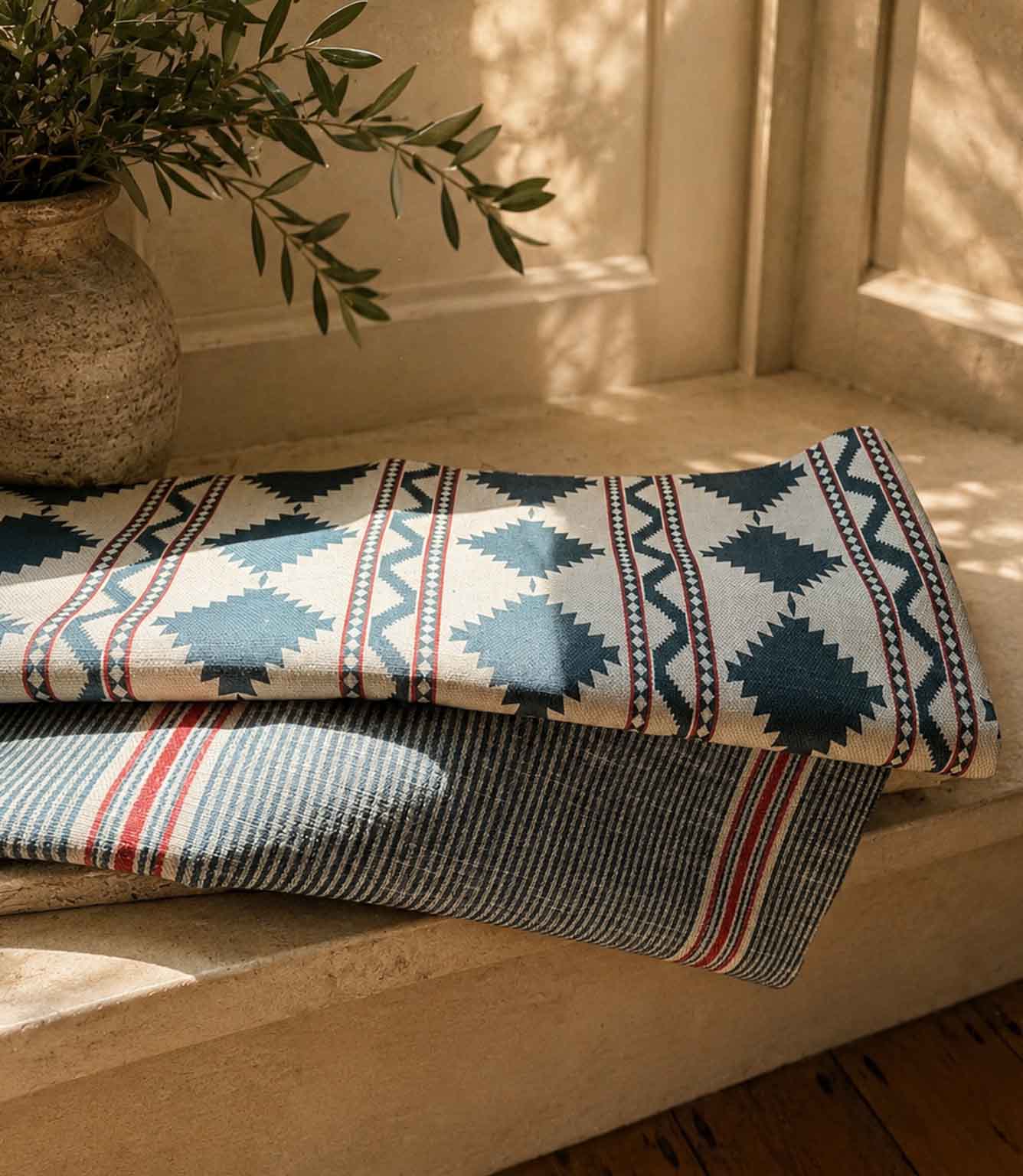 Patterned geometric and striped fabrics folded on a wooden surface with a plant in the background