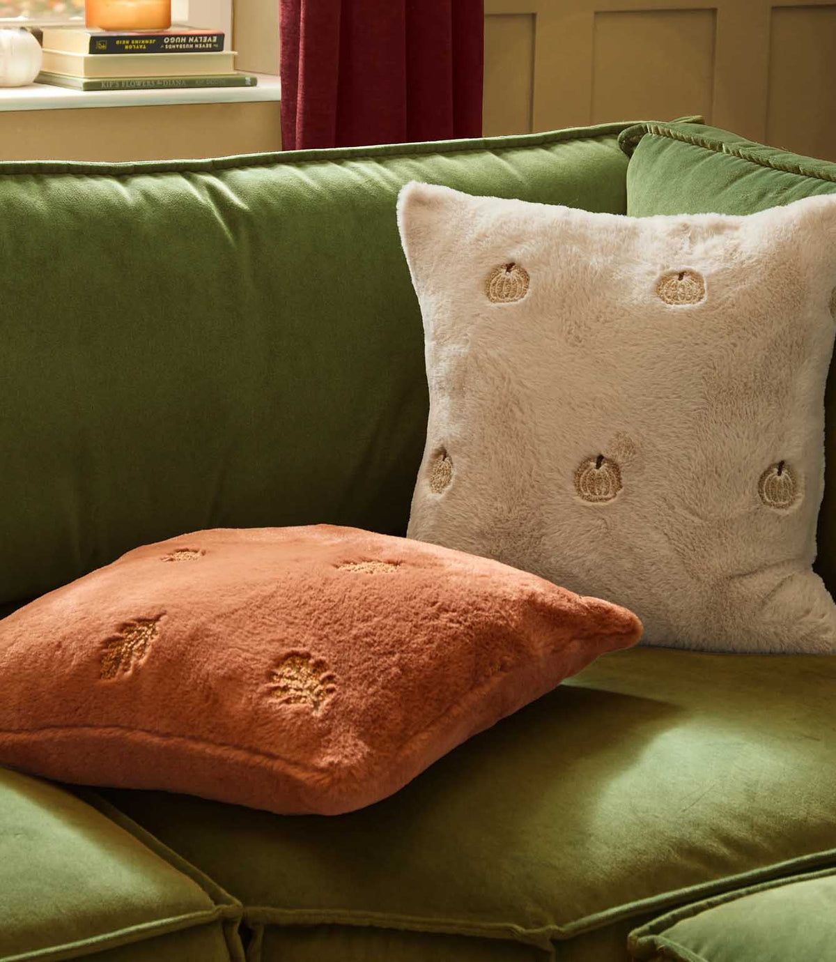 Natural cream pumpkin embroidered faux fur cushion styled with a rust orange pumpkin cushion on a green velvet sofa in an autumnal interior.