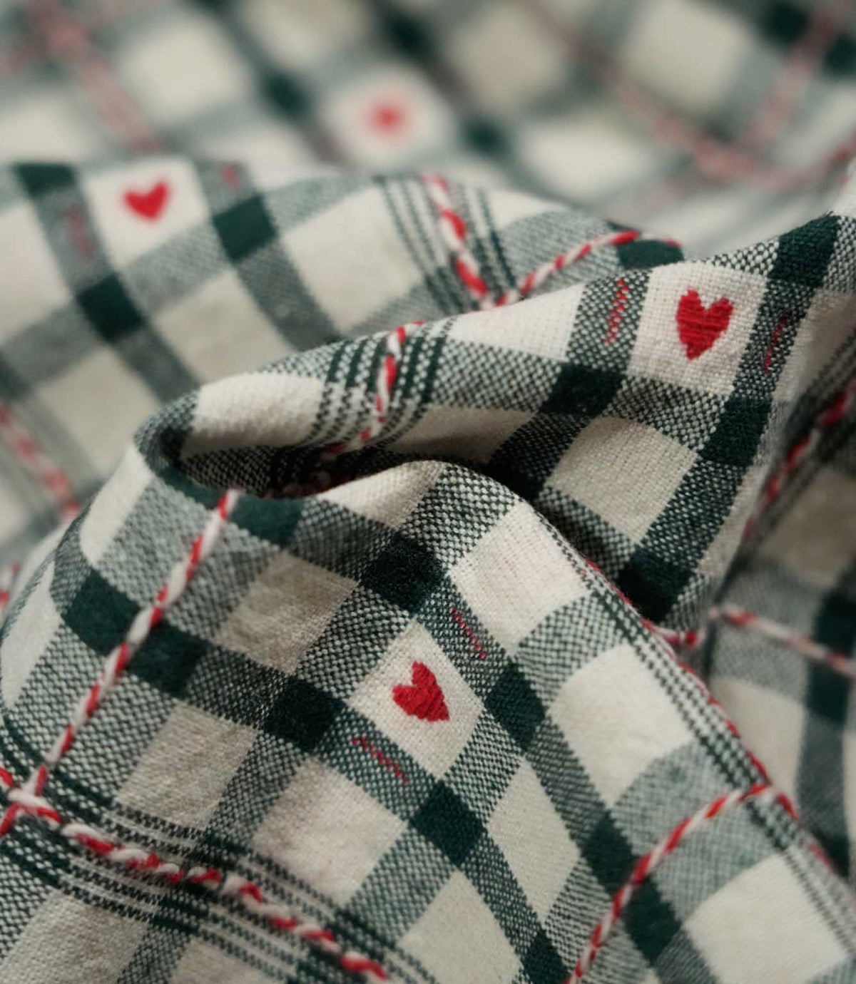 Macro close-up of Cotswold Heart Tartan fabric draped into soft folds, highlighting the woven green and ivory check, red hearts and contrasting thread details.