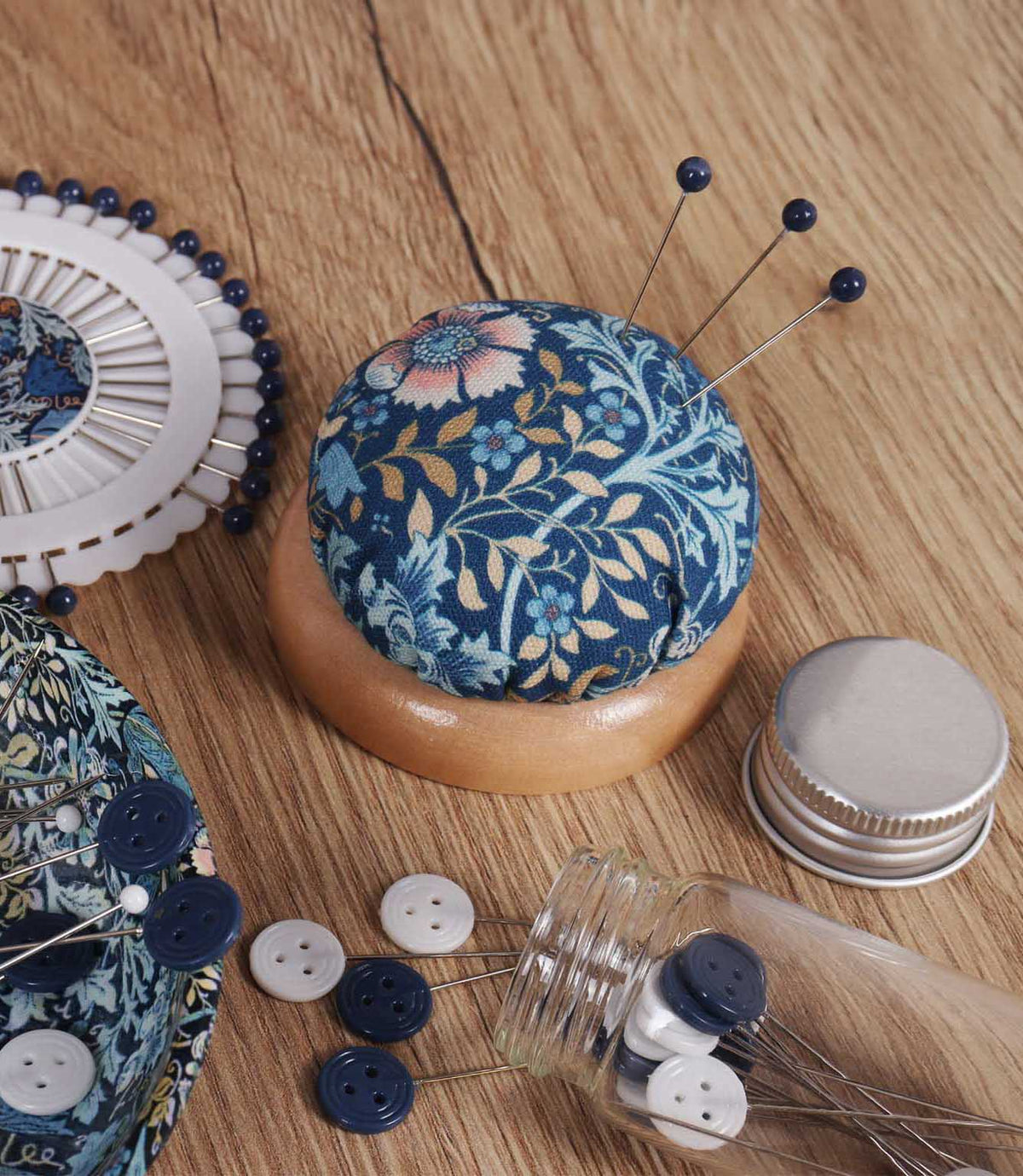 William Morris-inspired pincushion with a navy blue, turquoise, cream and gold floral fabric top and wooden base, holding three navy-headed sewing pins on a craft table.