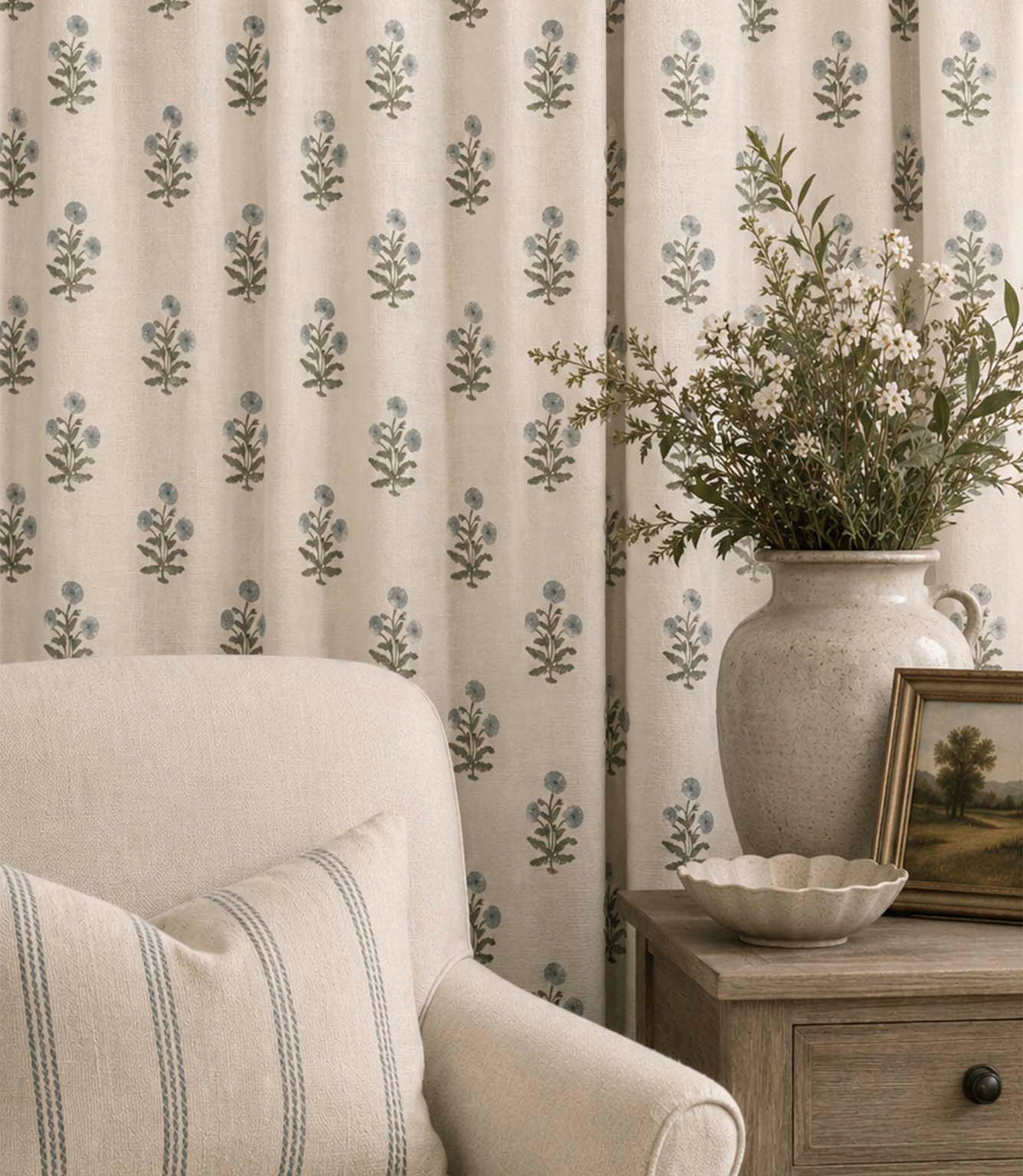 Novi Ink botanical print curtains with delicate blue flowers and green stems on a natural cream background, styled beside a neutral armchair and wooden side table with a ceramic vase of flowers.