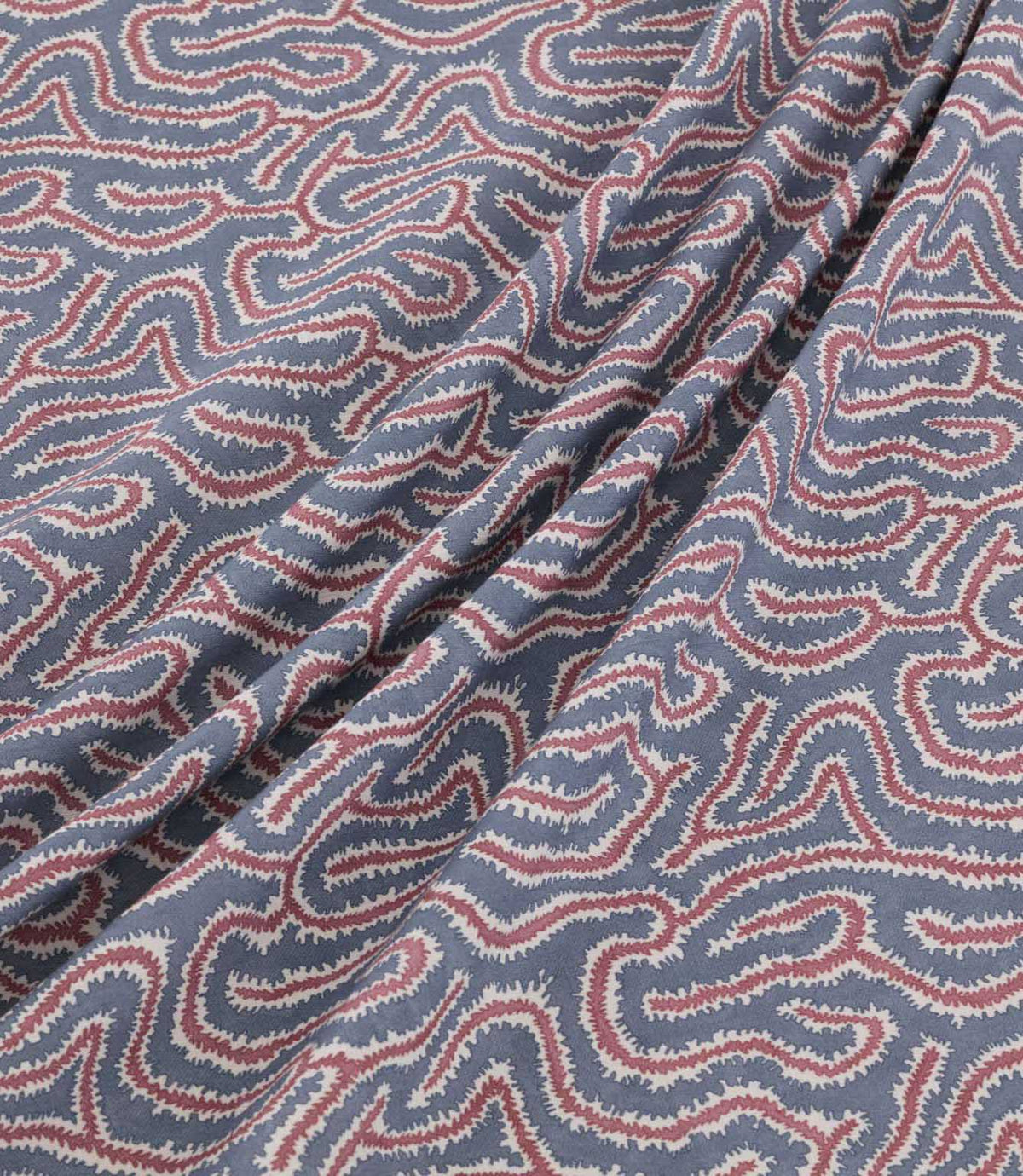 Noyack Anemone fabric arranged in soft, flowing folds, showcasing its slate blue background and repeating abstract pattern of winding dusty pink and ivory lines with delicate, feathered edges. The draped fabric highlights the intricate design and natural movement of the material.