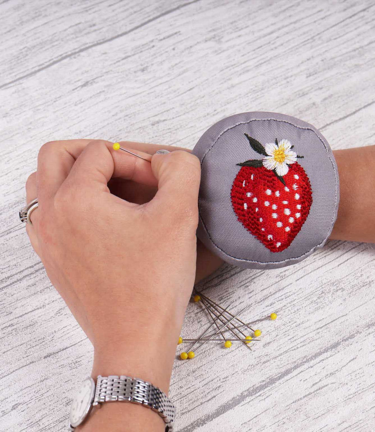 Grey wrist pincushion worn on the wrist while sewing, featuring an embroidered red strawberry with white flower and green leaves, with dressmaking pins nearby.