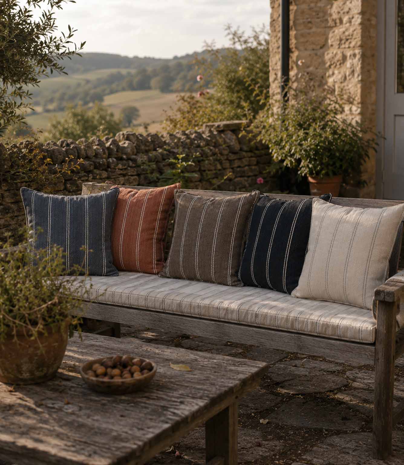 Outdoor setting with a wooden bench featuring striped outdoor cushions, stone wall, and greenery.