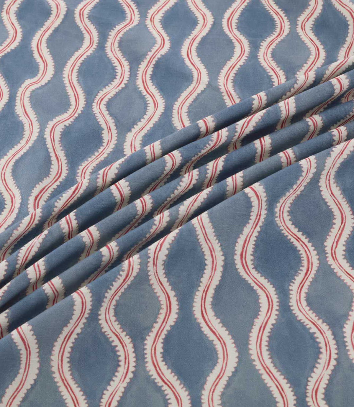 Wading Anemone fabric arranged in soft diagonal folds, showcasing its contemporary wave-inspired pattern in dusty blue, with cream scalloped edges, delicate red detailing and a subtly textured woven appearance.
