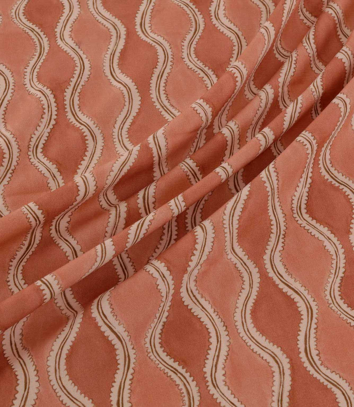 Wading Coralee fabric arranged in soft diagonal folds, showcasing its contemporary wave-inspired pattern in coral, terracotta and muted pink, with delicate cream scalloped edges, fine brown detailing and a subtly textured woven appearance.