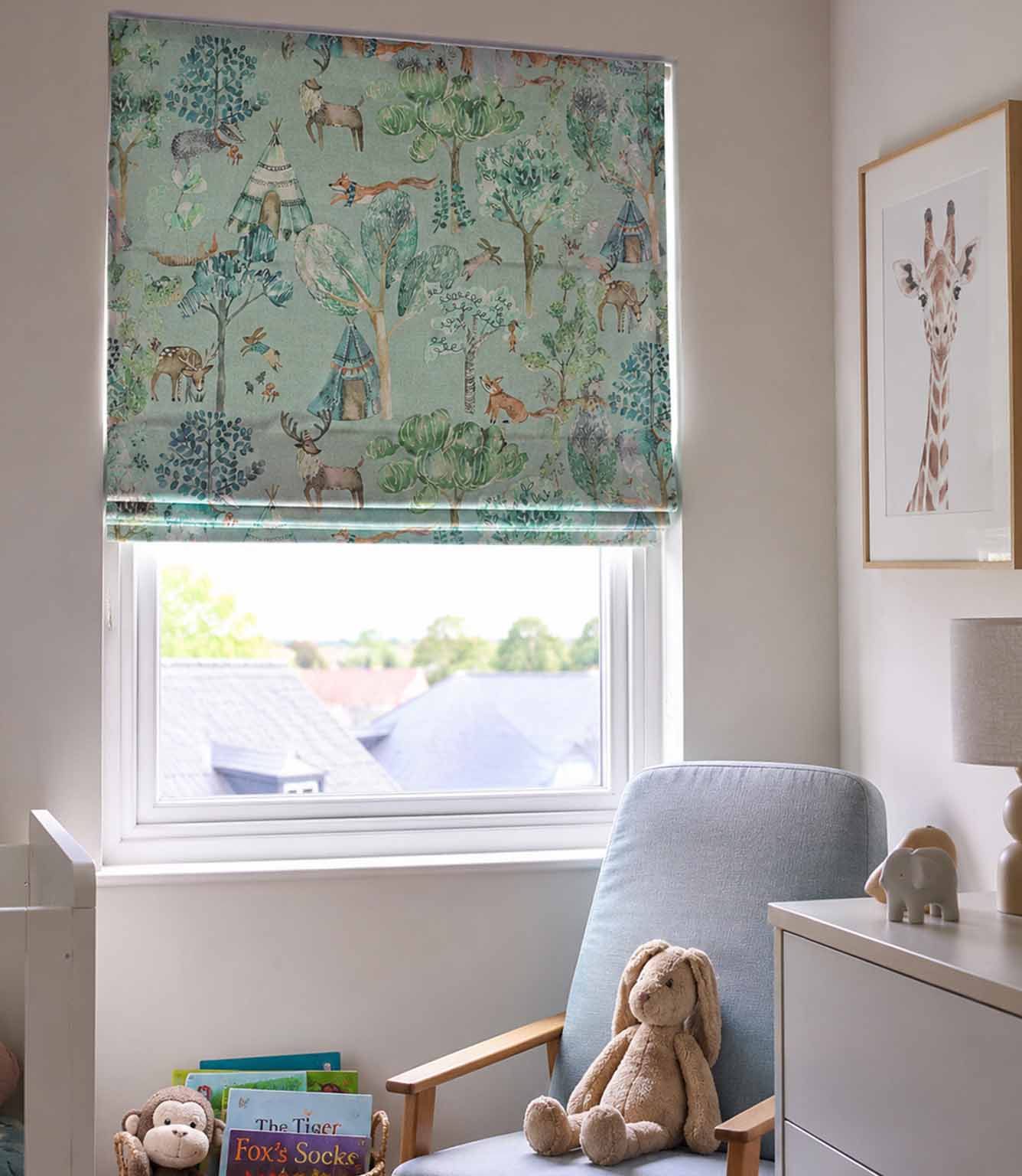 A window Roman blind with a woodland safari print featuring deer, foxes, and trees in muted green and blue tones, shown installed in a styled children's room with a blue armchair, stuffed bunny, and framed giraffe art.