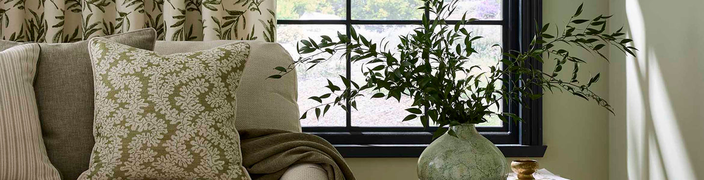 Neutral-toned living room with patterned cushions and curtains in soft green and cream, styled on a sofa beside a black-framed window with natural light and leafy greenery in a ceramic vase.