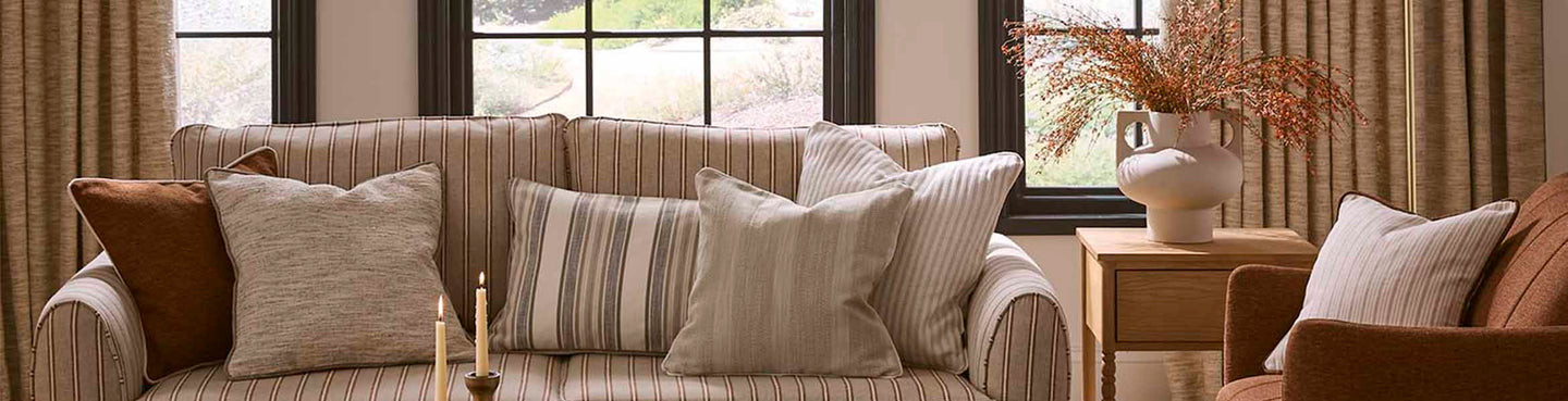 Neutral living room with a striped upholstered sofa, layered cushions in coordinating plains and stripes, and warm-toned curtains framing large windows.