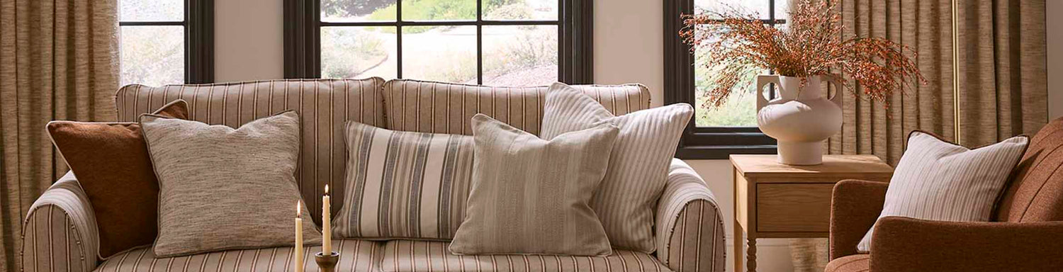 Neutral living room with a striped upholstered sofa, layered cushions in coordinating plains and stripes, and warm-toned curtains framing large windows.