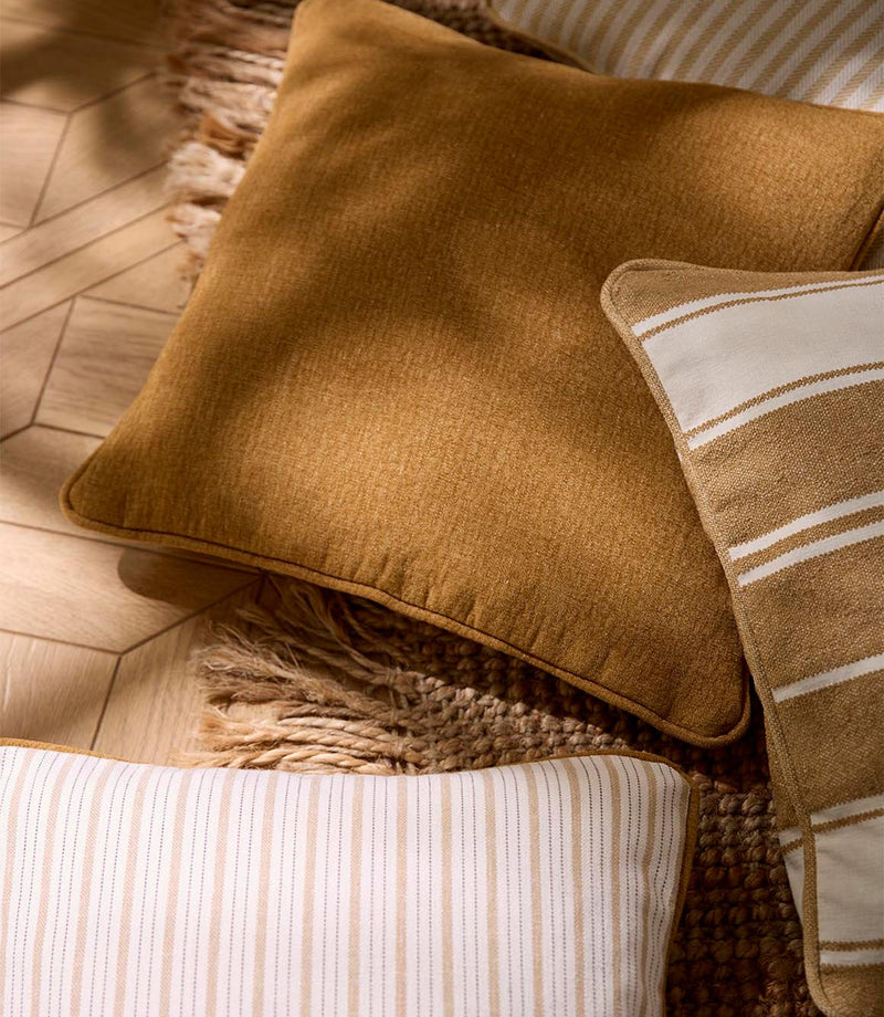 Warm-toned woven cushion arrangement featuring a plain textured fabric alongside striped designs in soft cream and golden ochre, styled on a wooden floor with natural fibres.