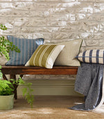 Rustic interior bench styled with woven striped cushions in soft blue, olive green and neutral tones, set against a textured stone wall with natural wood and greenery.