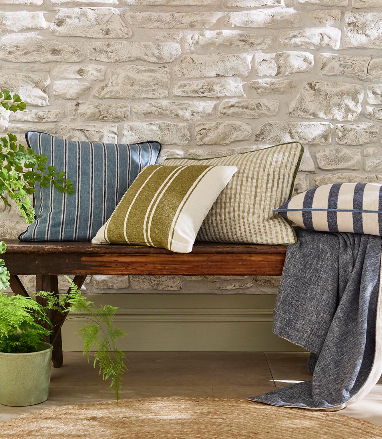 Rustic interior bench styled with woven striped cushions in soft blue, olive green and neutral tones, set against a textured stone wall with natural wood and greenery.