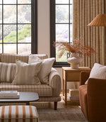 Bright living room with large windows, featuring a striped upholstered sofa and layered cushions in soft neutral and warm earthy tones, paired with textured curtains and natural wood furniture.