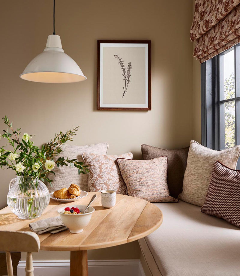 Dining nook with upholstered bench seating, layered cushions in textured woven and patterned fabrics, and a wooden table styled with flowers and tableware in warm neutral tones.
