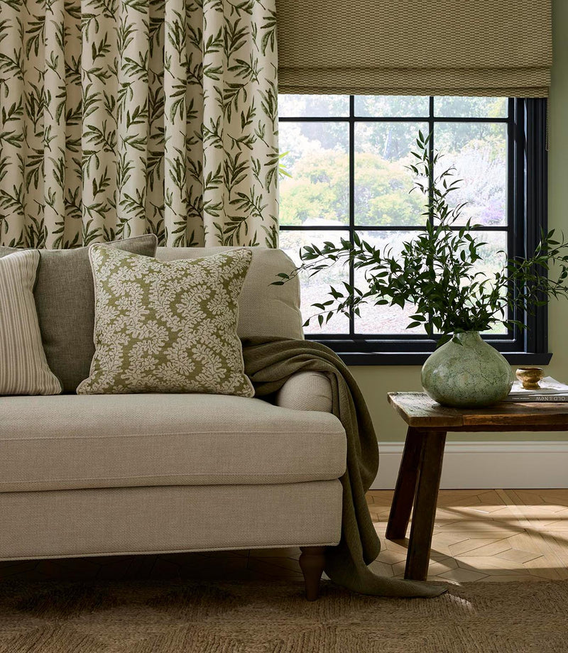 Living room scene featuring curtains in embroidered green leaf fabric, paired with a coordinating Roman blind, neutral sofa and patterned cushions for a calm, natural interior.