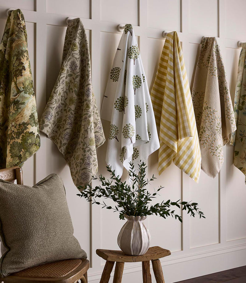 Selection of fabric samples draped on wall hooks, featuring botanical prints, trailing florals, stylised tree motifs and soft yellow gingham, styled above a wooden stool with a vase of greenery.