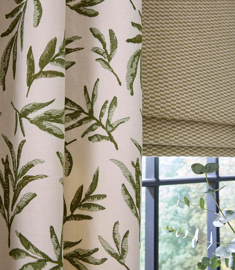Detailed view of embroidered green leaf curtain fabric alongside a small-scale geometric Roman blind, styled with a window and fresh greenery for a relaxed, cohesive look.