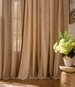 Light-filled interior scene with elegant striped curtains in soft neutral and warm golden tones, paired with sheer drapes and a wooden side table with fresh green flowers.