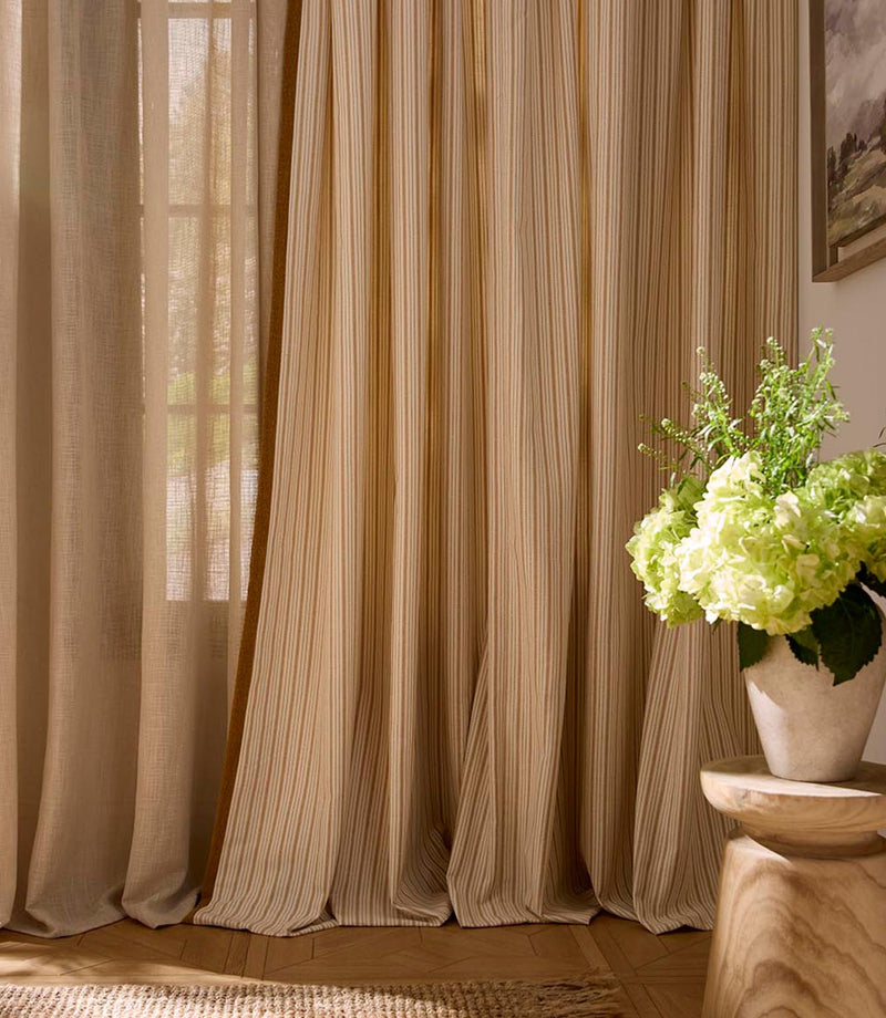 Light-filled interior scene with elegant striped curtains in soft neutral and warm golden tones, paired with sheer drapes and a wooden side table with fresh green flowers.