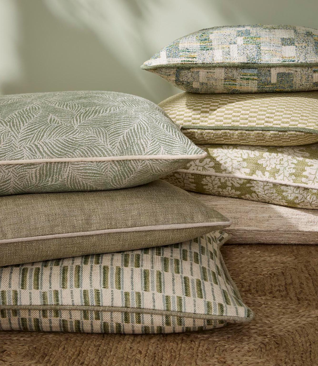 Styled arrangement of cushions in coordinating green and neutral fabrics, including geometric, leaf and textured weaves, layered on a natural jute rug.