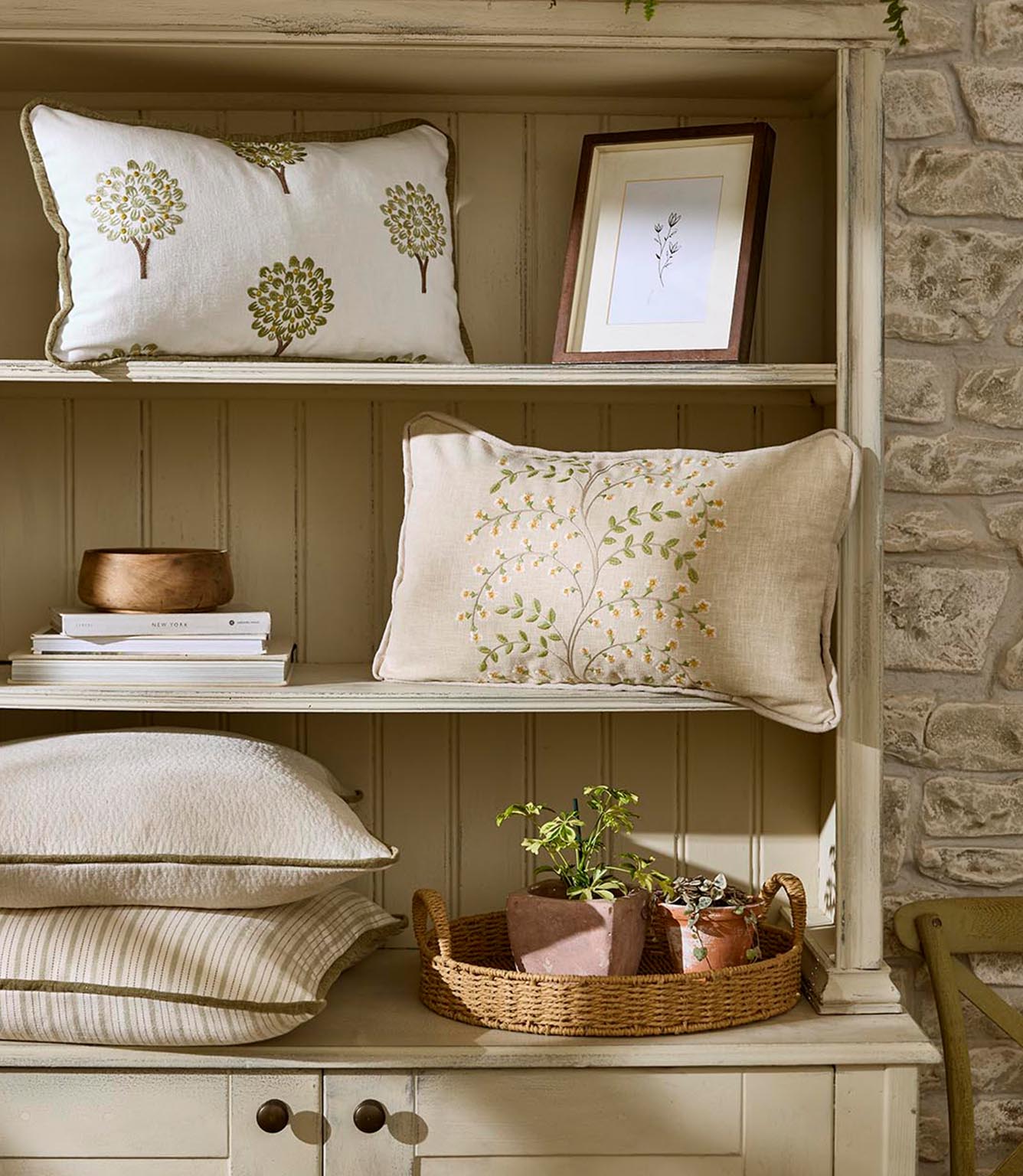 Painted wooden shelving styled with cushions in embroidered floral and stylised tree fabrics, alongside stacked books, ceramics and potted plants for a relaxed, natural interior look.