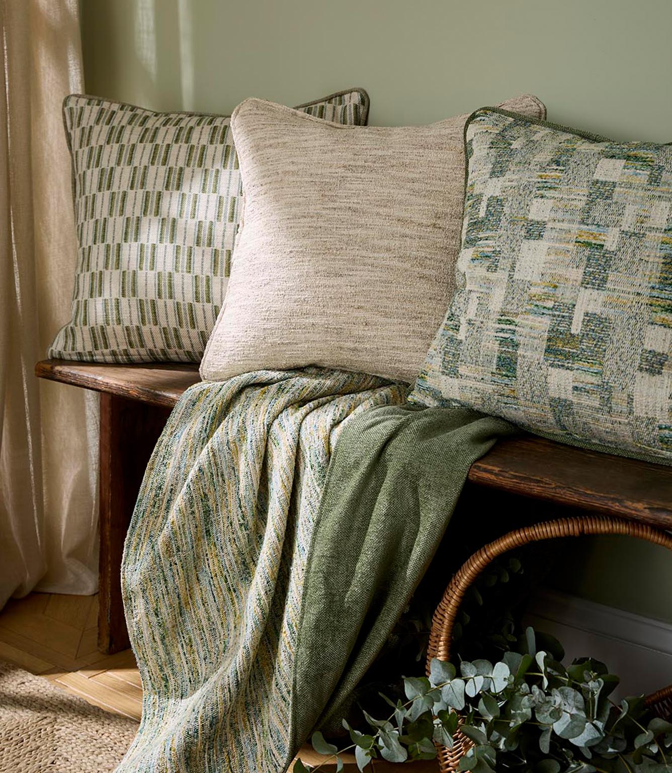 Interior setting with cushions in soft green and neutral upholstery fabrics on a wooden bench, paired with a draped throw and natural accessories for a relaxed, contemporary look.