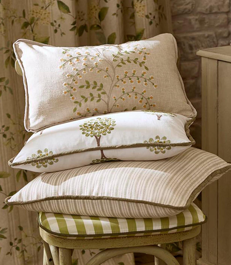 Layered cushions in complementary fabrics, including embroidered florals, tree motifs and fine ticking stripes, stacked on a wooden stool in front of coordinating botanical curtains.