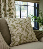 Styled interior scene showing a cushion upholstered in olive green and off-white foliage fabric, placed on a neutral sofa beside a window with soft natural light and coordinating botanical accents.