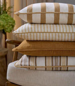 Stack of upholstered cushions in coordinating woven fabrics, combining plain and striped designs in warm ochre and soft neutral tones, creating a layered, relaxed look.