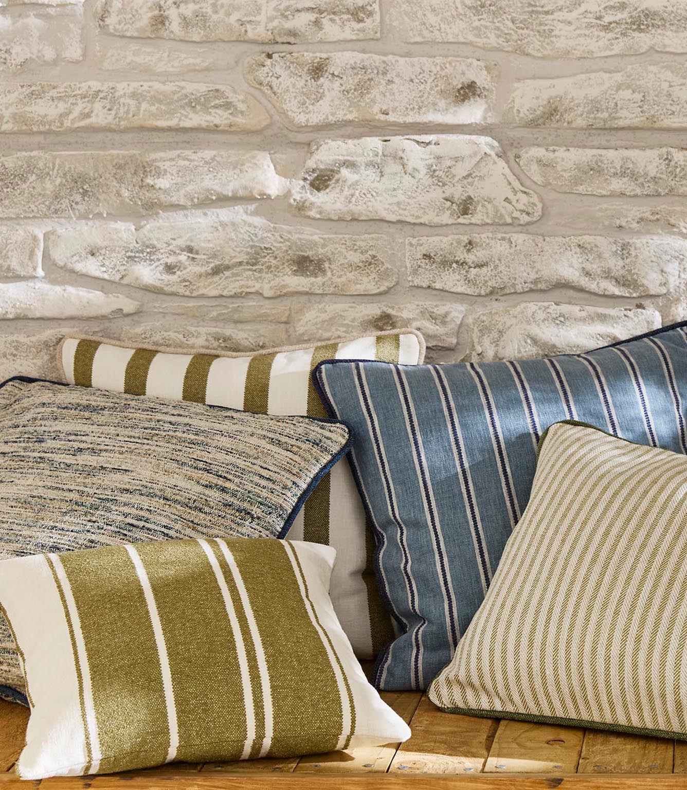 Close-up arrangement of layered cushions in mixed woven stripes and textured plains, featuring soft blue, olive green and neutral shades on a wooden bench for a relaxed, coordinated look.