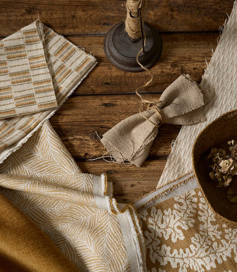 Flat lay of coordinated upholstery fabrics in warm neutral and ochre tones, showing a mix of woven textures and patterns on a rustic wooden surface with styling accessories.