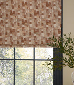 Roman blind in a woven geometric fabric with an abstract check pattern in warm rust and natural linen tones, styled over a black-framed window with soft daylight and greenery visible outside.