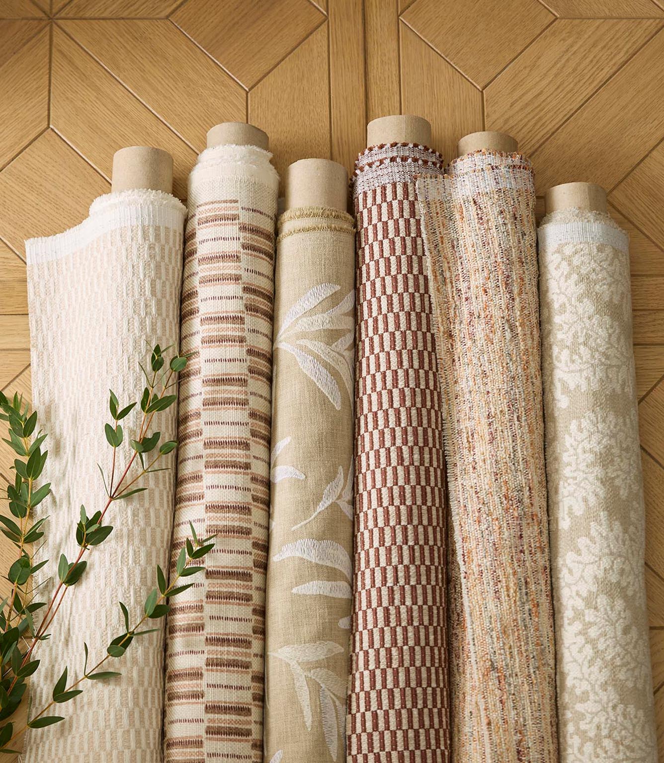Collection of rolled upholstery fabrics in complementary natural tones, including stripes, checks and textured weaves, displayed against a wood backdrop.