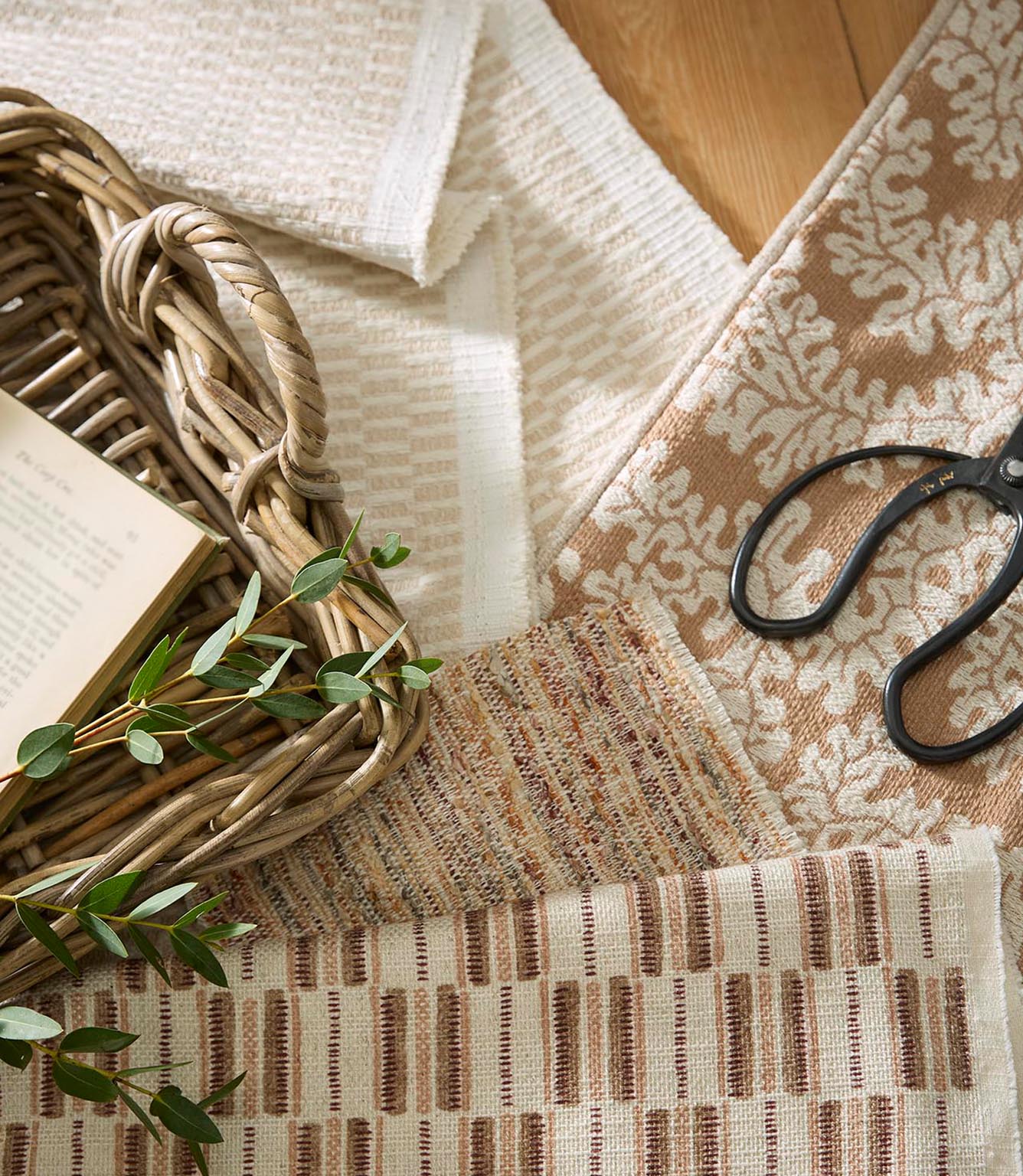 Fabric samples arranged with a woven basket, scissors and greenery, showing coordinating neutral upholstery fabrics with stripe and textured patterns.
