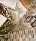 Close-up of layered fabric samples including a textured woven upholstery fabric in warm beige and rust tones, alongside patterned textiles, a woven basket and tailoring scissors.