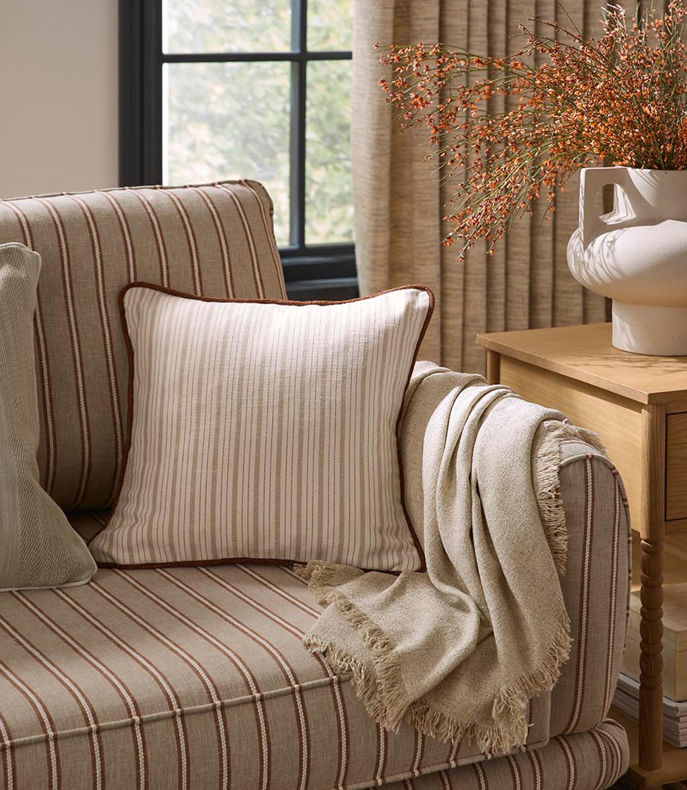 Striped upholstered armchair with coordinating cushion and soft throw, styled in warm neutral tones alongside woven curtains and natural wood accents.