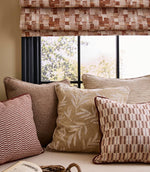 Interior scene with layered cushions in coordinating fabrics, including embroidered floral, geometric weave and textured plains in warm neutral and earthy brown tones.