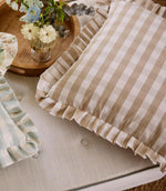 Pair of ruffle-edged cushions in soft beige gingham and pale blue stripe fabrics, arranged on a neutral upholstered seat with a wooden tray of fresh flowers.