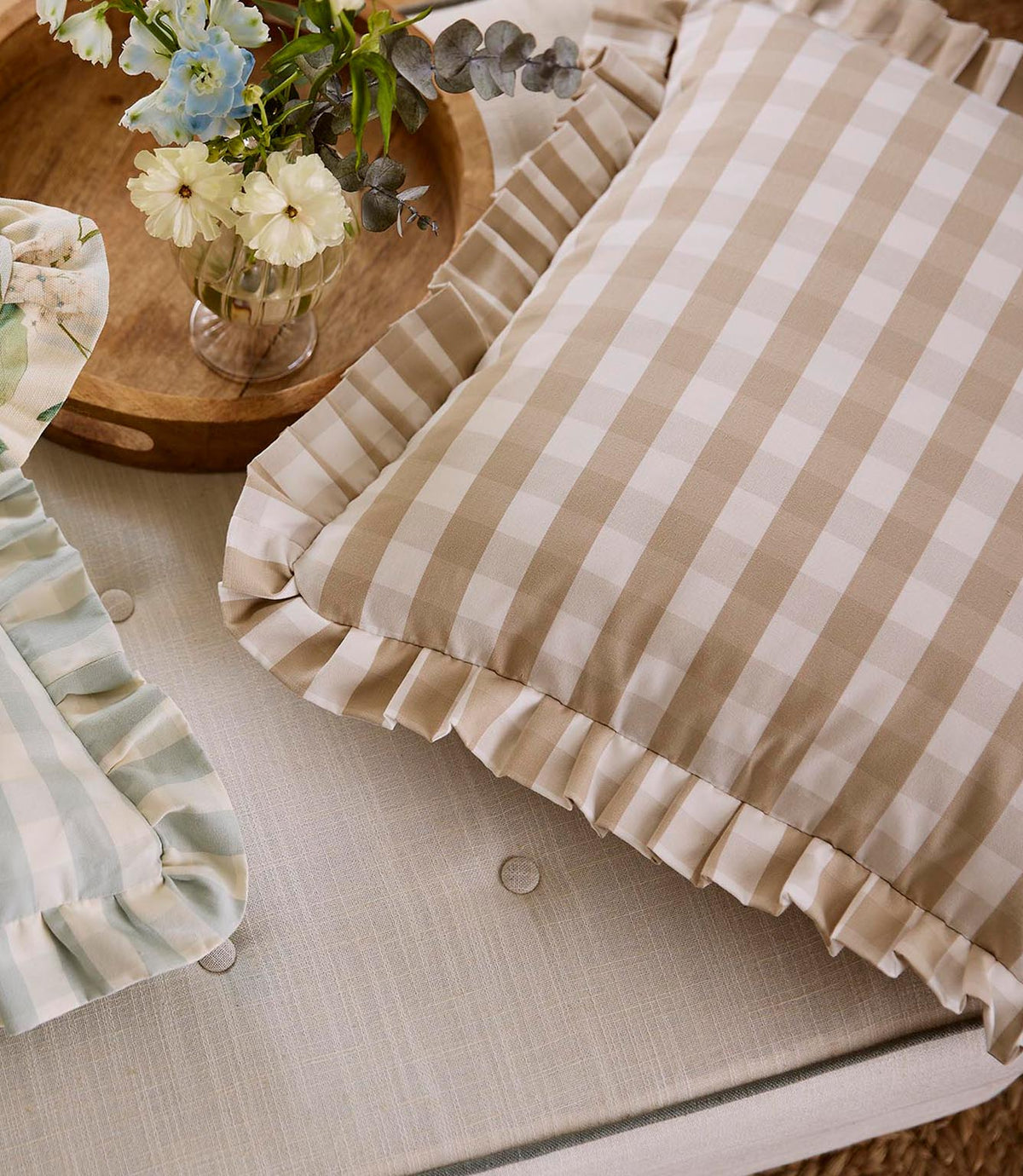 Pair of ruffle-edged cushions in soft beige gingham and pale blue stripe fabrics, arranged on a neutral upholstered seat with a wooden tray of fresh flowers.