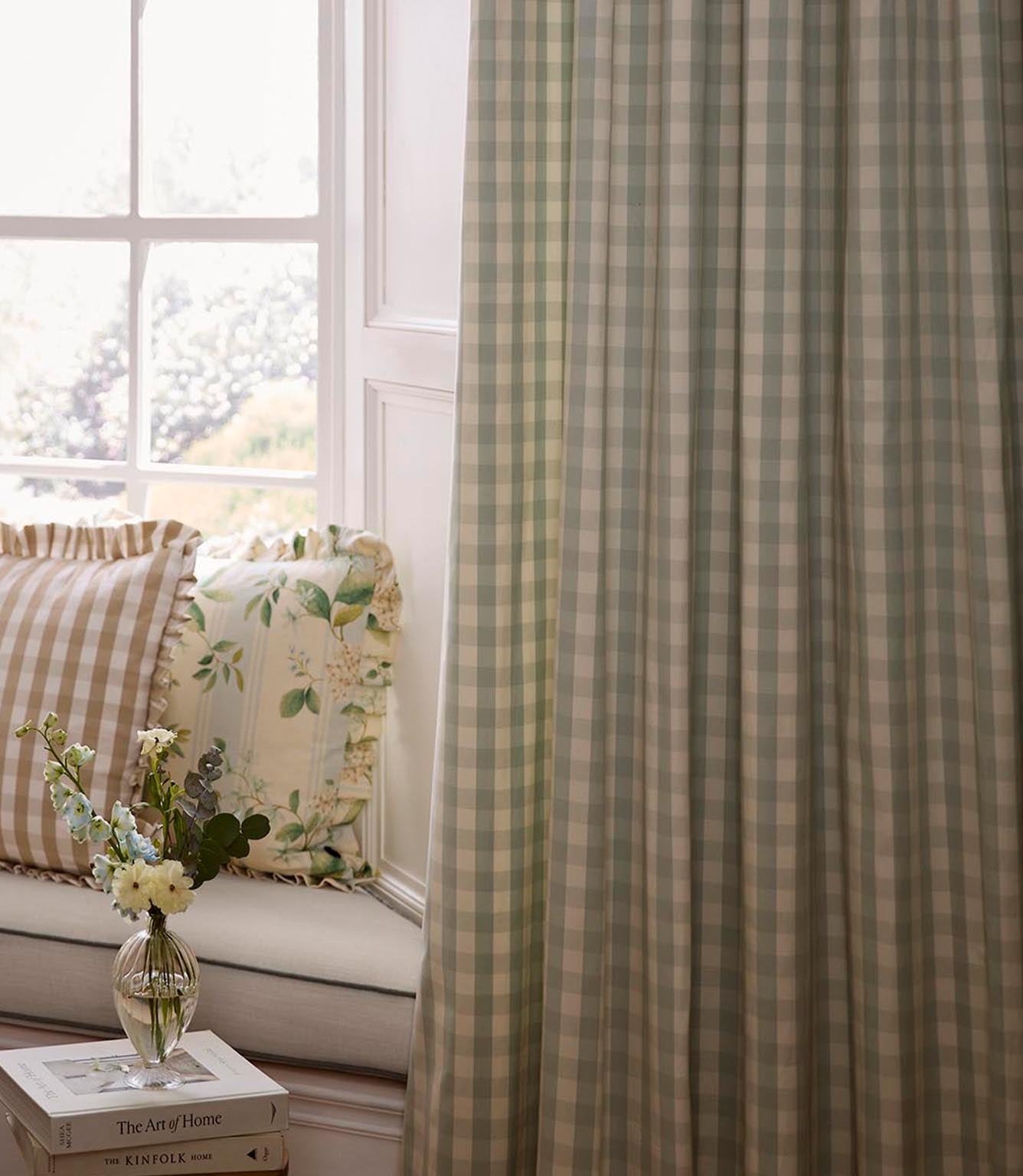 Window seat styled with layered cushions in gingham and botanical prints, set beside full-length green check curtains and a small vase of flowers on stacked books.