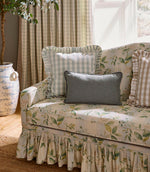 Floral upholstered sofa with a gathered skirt, styled with gingham and plain cushions, set against soft green check curtains in a bright, relaxed living space.