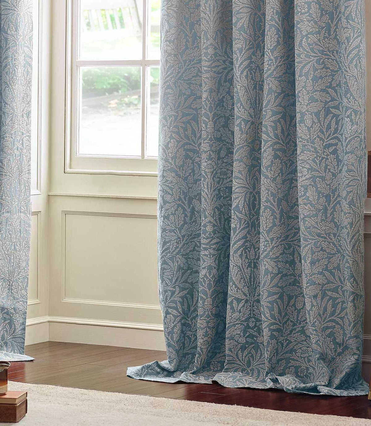Full-length Morris & Co Acorn jacquard curtain in Wedgwood blue and off-white, draping onto a wooden floor beside a traditional panelled window.