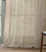 Full-length Morris & Co Acorn sheer curtain in cream and beige, softly draping onto a wooden floor beside a pale green framed window.