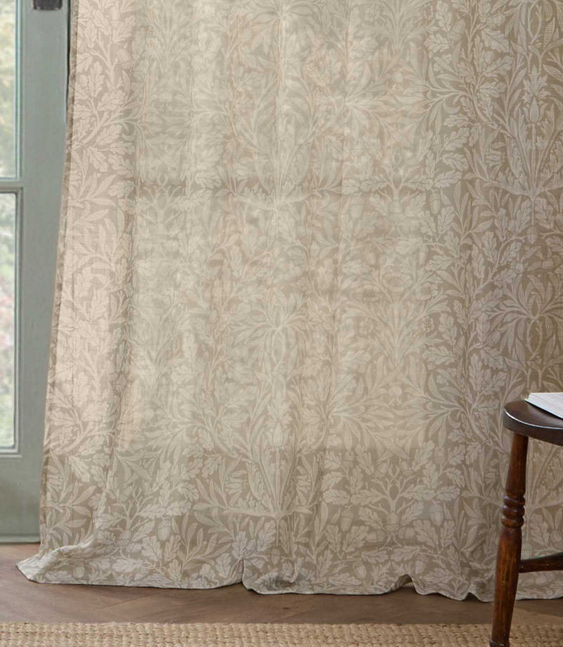 Full-length Morris & Co Acorn sheer curtain in cream and beige, softly draping onto a wooden floor beside a pale green framed window.