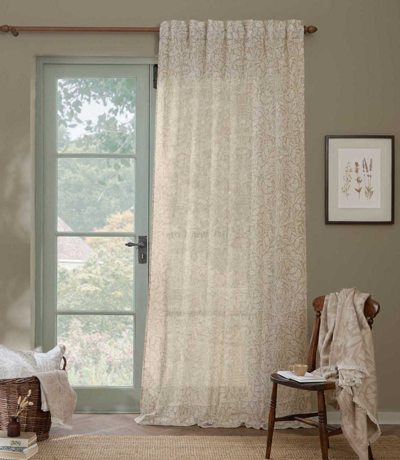 Morris & Co Acorn sheer curtain in cream and beige hanging beside a pale green glazed door, styled with a wooden chair and natural woven accessories.
