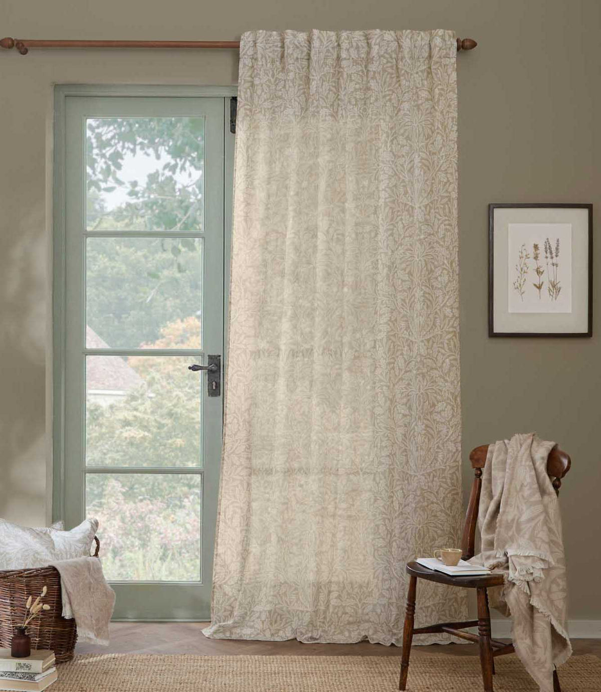 Morris & Co Acorn sheer curtain in cream and beige hanging beside a pale green glazed door, styled with a wooden chair and natural woven accessories.